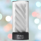 A white spiral-patterned cylindrical object, the TENGA 3D Spiral Penis Masturbator from Tenga, is encased in a clear holder with a black base labeled "SPIRAL TENGA 3D." Made from antibacterial elastomer, it is set against a softly blurred backdrop of multicolored lights.